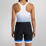 Rear view of a woman wearing a sleeveless athletic one-piece unisuit with a white upper back, blue wavy stripe pattern across the waist, and black shorts with blue and white leg bands, set against a light gray studio background.