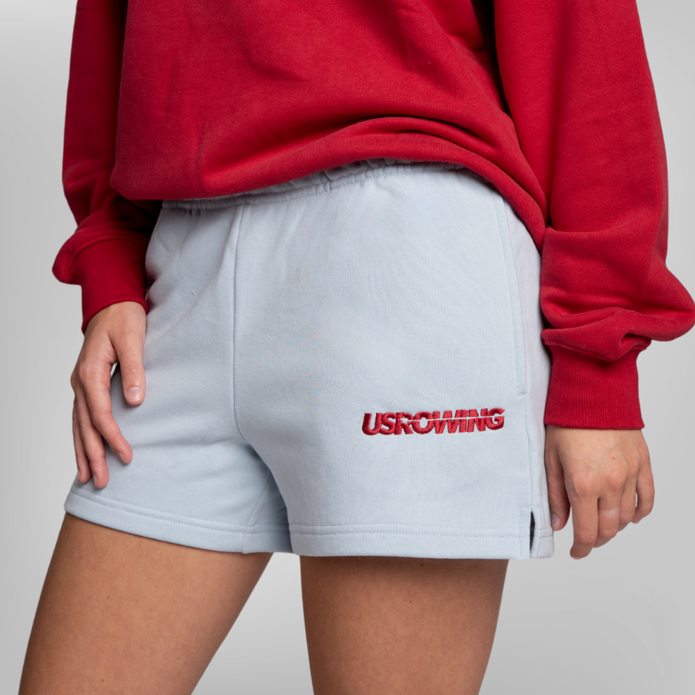 A person wearing light blue sweat shorts with a red USRowing logo on the left leg, paired with a maroon sweatshirt.