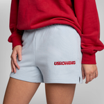 A person wearing light blue sweat shorts with a red USRowing logo on the left leg, paired with a maroon sweatshirt.