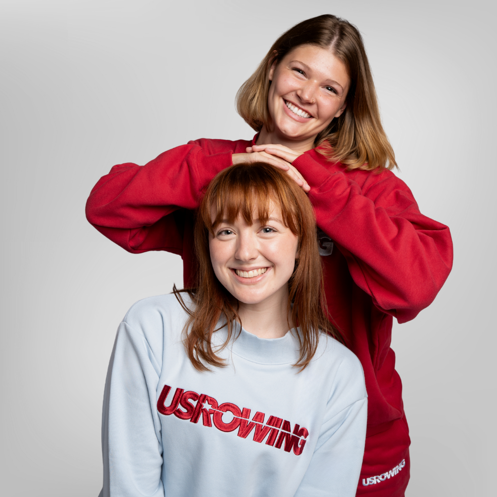 Two modelsposing together against a light gray studio background. The woman in the back wears a maroon sweatshirt, and the woman in the front wears a light blue sweatshirt with 'USROWING' embroidered across the chest in red.