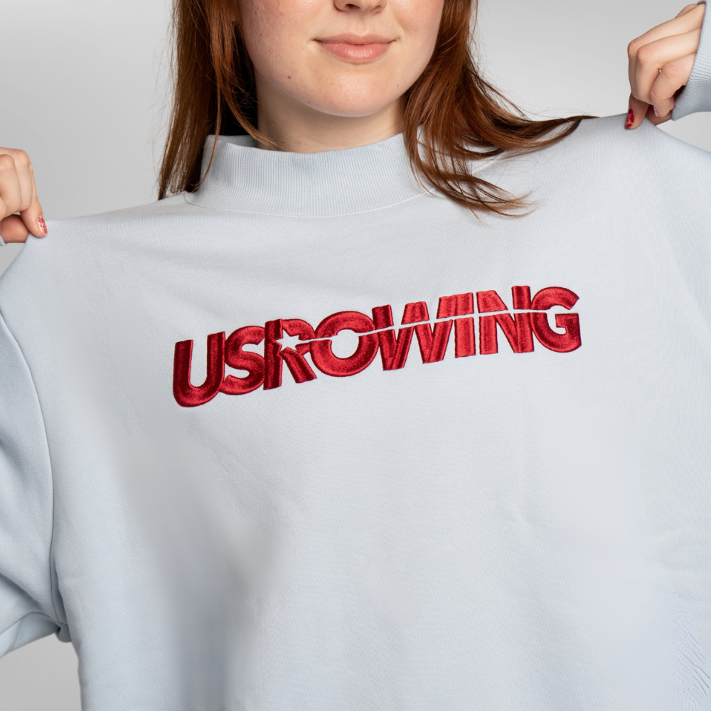 A close-up of a person wearing a light blue mock-neck sweatshirt with the word 'USROWING' embroidered across the chest.