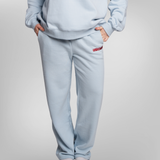 A person wearing a light blue sweatshirt and matching sweatpants with a small red embroidered USRowing logo on the left hip, shown against a plain light gray studio background.