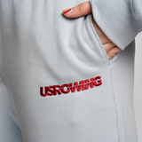 Close-up of a person wearing a light gray sweatshirt, showing a red embroidered USROWING logo on the leg.