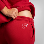 A close-up of a person wearing a red sweatshirt and matching red sweatpants, lifting the sweatshirt to show the side pocket and white embroidered star detail on the pants.