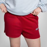 A close-up of a person wearing maroon sweat shorts with a small white embroidered logo on the lower left leg, paired with a light blue sweatshirt.