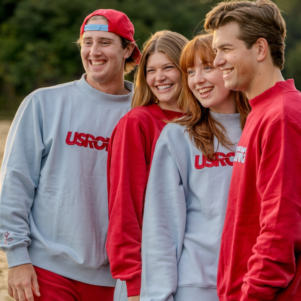 Four smiling young adults standing outdoors in matching USRowing mockneck sweatshirts in red and light gray, with red sweatpants and a red cap.