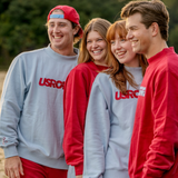 Four smiling young adults standing outdoors in matching USRowing mockneck sweatshirts in red and light gray, with red sweatpants and a red cap.
