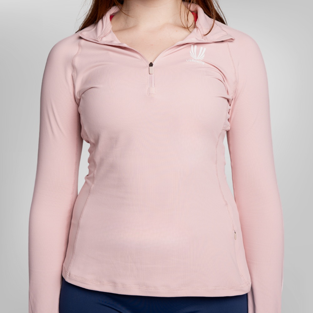A woman wearing a light pink long-sleeve athletic quarter-zip pullover with a small USRowing logo on the left chest, shown against a neutral light gray background.